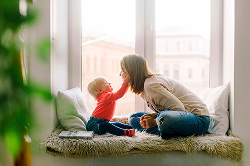 Toddler touching a mom's face while sitting on a window seat with sun coming in the window.